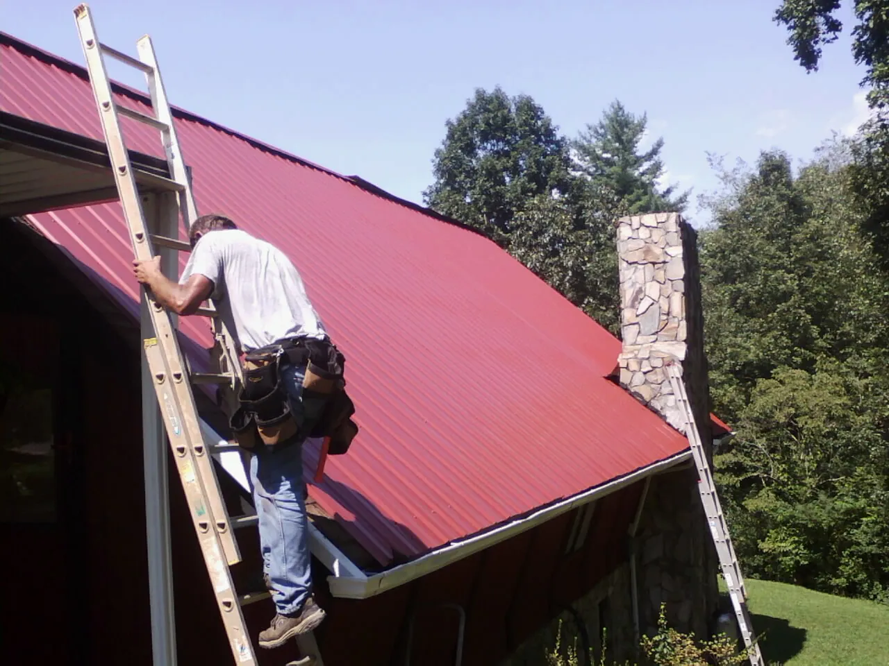 Roofer on a ladder beside a red metal roof and stone chimney — Soffit & Fascia Repair by Roofing Brothers in Paskenta